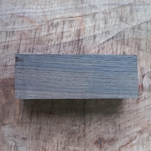 May include: A close-up of a dark gray wooden block with a wood grain pattern. The block is sitting on a light brown wooden surface.