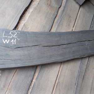 Op de afbeelding: Een donkergrijze houten plank met een natuurlijke scheur die door het midden loopt. De plank is 52 inch lang en 11 inch breed. De afmetingen zijn met witte krijt op het hout geschreven.