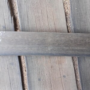 May include: A rectangular, grey wooden plank, approximately 63.5 cm long and 12.7 cm wide, lies on a weathered wooden surface. The plank's surface has a visible wood grain texture. White numbers indicate the dimensions of the plank.