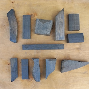 May include: A collection of eleven dark grey, weathered wooden blocks and offcuts of various shapes and sizes, including rectangular, triangular, and irregular forms, displaying visible wood grain textures and natural edges on a light wood surface.