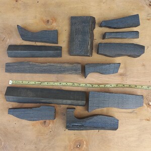 May include: A collection of eleven dark grey, weathered wooden blocks of varying shapes and sizes arranged on a light wood surface. A yellow measuring tape is positioned horizontally across the centre, showing a length of approximately 60 centimetres.