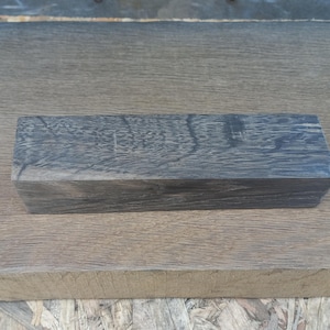 May include: A rectangular block of dark gray wood with visible grain patterns. The block is positioned on a light brown wooden surface, showcasing the contrast in textures and colors. The wood appears to be unfinished.