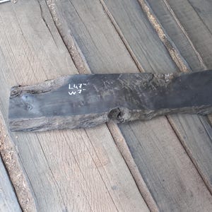 May include: A dark gray, weathered wooden plank with the text "L47" W7" in white. The plank is approximately 47 inches long and 7 inches wide. The wood has a rough, natural edge and is set against a background of wooden planks.