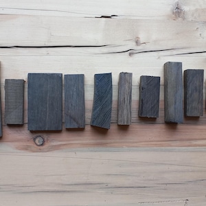 May include: An overhead shot displays a collection of ten rectangular wooden blocks in varying sizes and shades of gray. The blocks are arranged in a row on a light brown wooden surface, showcasing the natural wood grain.