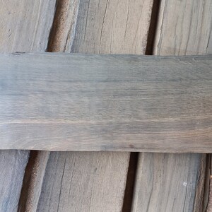May include: A weathered gray wooden plank with a rough, uneven edge. The plank has a natural wood grain pattern and is lying on a surface of similar wood planks.