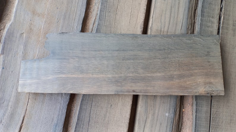 May include: A weathered gray wooden plank with a rough, uneven edge. The plank has a natural wood grain pattern and is lying on a surface of similar wood planks.