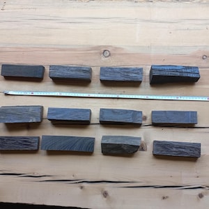 May include: An overhead shot shows twelve rectangular, dark gray wooden blocks arranged on a light brown wooden surface. A measuring tape is placed across the blocks. The blocks appear to be roughly 4 inches long.