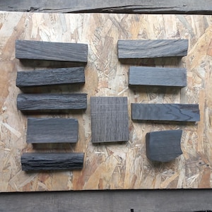 May include: A collection of dark gray wooden blocks and a square piece of wood arranged on a light brown particle board. The wood pieces vary in shape and texture, showcasing different grain patterns and shades of gray.