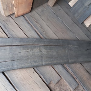 May include: A dark grey wooden plank, showing signs of weathering and cracks, with white text indicating dimensions. It is surrounded by other wooden pieces, suggesting a lumber or woodworking context.