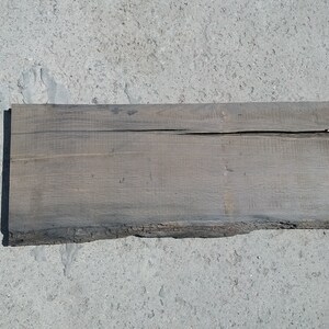 Op de afbeelding: Een verweerd, grijs houten plank met een ruwe, natuurlijke rand. De plank heeft een scheur die door het midden loopt.