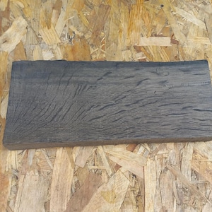 May include: A rectangular, dark gray wooden plank with visible wood grain. The plank has a rough, textured surface and is displayed against a background of wood chips.