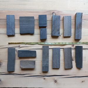 May include: An overhead shot of various gray wooden blocks of different shapes and sizes, arranged on a light brown wooden surface. A yellow measuring tape is laid across the blocks. The blocks appear to be for a woodworking project.