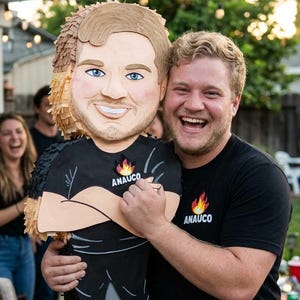 Peut inclure: Une piñata personnalisée en forme de personne aux cheveux châtain clair, portant un t-shirt noir avec un motif de flamme et le texte ANAUCO. La piñata présente des détails en papier frangé, un visage souriant et un pantalon noir avec des baskets.