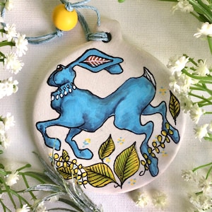 May include: A blue ceramic ornament with a painted image of a blue rabbit with a white pearl necklace, surrounded by yellow and green leaves.