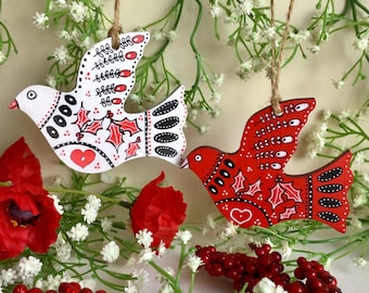 Könnte beinhalten: Zwei hölzerne Tauben-Ornamente, eine weiß mit roten Akzenten und eine rot mit weißen Akzenten. Beide Tauben haben ein Herz- und Stechpalmenblatt-Design.