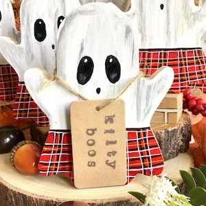 May include: White wooden ghost painted with black eyes and a red and black plaid skirt. The ghost is holding a brown tag that says "Kilty Boo's".