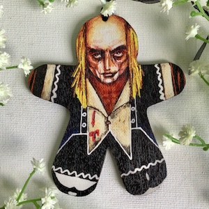 Puede incluir: Un adorno de madera de hombre de jengibre con una imagen pintada de una persona con cabello largo y rubio, vistiendo un vestido blanco y negro con un cuello blanco y una mancha roja en el pecho. La persona tiene una tez pálida y una expresión seria.