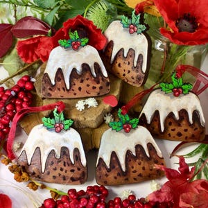 Pode incluir: Enfeites de Natal de madeira com formato de pudim de Natal. Pintados com glacê branco, base marrom e detalhes de azevinho. Possuem fitas vermelhas para pendurar. A cena inclui bagas e flores vermelhas, evocando um ambiente festivo.
