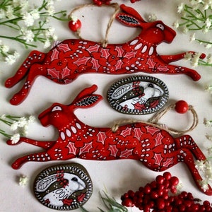 Peut inclure: Deux ornements en bois rouge en forme de lièvre, avec des motifs de feuilles de houx blancs et verts et une pose de course. Deux pierres peintes de forme ovale avec un motif de lapin blanc sont également présentes. Les ornements sont attachés avec de la ficelle et des perles rouges.