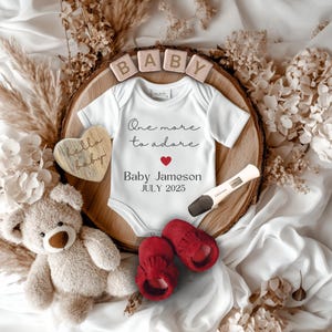 May include: A baby announcement featuring a white onesie with the text "One more to adore" and "Baby Jameson JULY 2025". The announcement includes a wooden heart, red baby shoes, a teddy bear, and a pregnancy test.