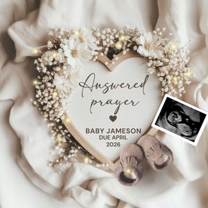 Könnte beinhalten: Ein herzförmiger Holzrahmen mit den Worten "Answered prayer" und "Baby Jameson Due April 2026". Der Rahmen ist mit weißen Blumen und Lichterketten verziert. Ein Ultraschallbild und Babyschuhe sind ebenfalls enthalten.