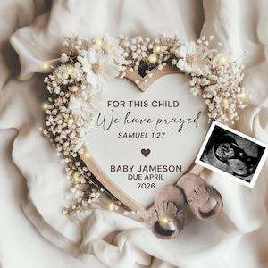 May include: A heart-shaped wooden frame with the text "For this child we have prayed" and "Baby Jameson Due April 2026". The frame is decorated with white flowers and fairy lights. A sonogram image and baby shoes are also included.