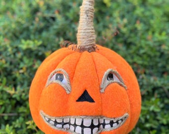 May include: Orange felt pumpkin decoration with a smiling face, featuring a black triangle nose and a wide, toothy grin. The eyes are detailed with twine accents. The stem is made of twisted rope. Perfect for Halloween.