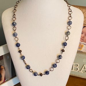 May include: A necklace with blue and white speckled beads and copper-colored chain links. The necklace is displayed on a white mannequin bust. The beads are round and evenly spaced along the chain, creating a simple yet elegant design.