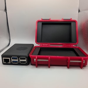 May include: A black electronic device with multiple USB ports and an Ethernet port sits next to an open red case. The case has a black interior and metal latches. The case is designed to protect the electronic device.