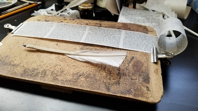 May include: A white feather quill pen rests on a wooden writing surface. A long strip of paper with handwritten text is clipped to the surface. The paper is rolled up at the end.