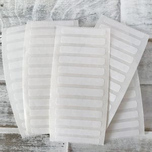 May include: Several sheets of white, rectangular labels with rounded corners are fanned out against a light wood-grain background. Each label sheet has multiple rows of individual labels, ideal for organizing or labeling items.