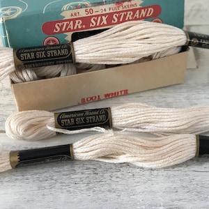 May include: A vintage cardboard box labeled Star Six Strand containing off-white embroidery cotton skeins. Each skein features a black and gold paper band with the text American Thread Co. Star Six Strand Embroidery Cotton printed on it.