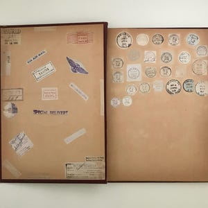 May include: An open vintage scrapbook with a brown cover and pages. The left page features various postage stamps and labels, including "VIA AIR MAIL" and "SPECIAL DELIVERY." The right page is filled with circular stamps.