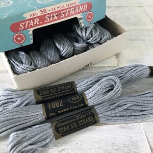 May include: A box of gray embroidery floss labeled "STAR SIX STRAND" with the text "ART. 50-24 PULL SKEINS." Three skeins of gray floss are in the foreground, each with a black label that reads "1062 FAST COLOR." The box is light blue and white.
