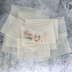 May include: A collection of translucent, off-white glassine envelopes stacked in an overlapping arrangement. One envelope in the center contains a vintage postage stamp featuring two American flags and the text United States 10 cents.