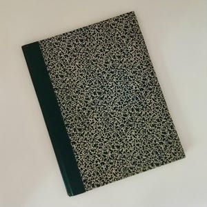May include: A vintage composition notebook with a black and white speckled cover and a dark green spine. The notebook is closed and lies flat on a white surface. The cover pattern is reminiscent of a classic design.