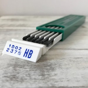 May include: A green and white pencil lead container with the text "1 DOZ 2375 HB" in blue. The container is open, revealing several dark gray pencil leads.