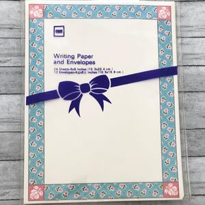 May include: A package of writing paper and envelopes. The package is wrapped with a dark blue ribbon and bow. The border is light blue with a floral pattern. The text on the package reads "Writing Paper and Envelopes". The paper measures 6x8 inches.