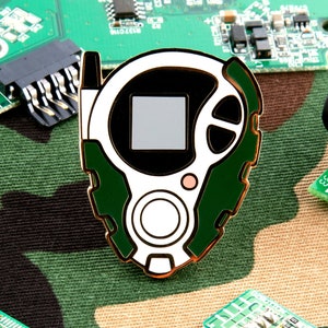 May include: A green and white enamel pin of a digital device with a screen and buttons. The pin is on a camouflage background with circuit boards.