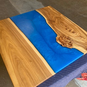 May include: A wooden table top with a blue epoxy river running through the center. The wood has a natural grain pattern and the epoxy is smooth and shiny.