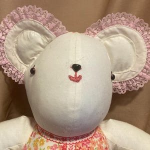 May include: A white teddy bear with pink lace trim on the ears. The bear has black button eyes and a red embroidered nose and mouth. The bear is wearing a floral print dress.