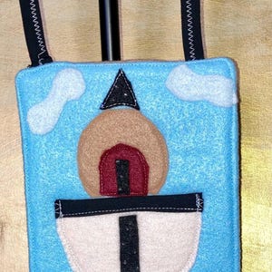 May include: A rectangular light blue felt bag featuring a layered applique design of a rocket ship launching into a sky with white clouds. The rocket has a tan body, a red window, and a black base with red flames. It includes two black straps.