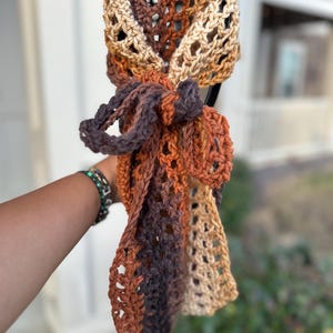 May include: A hand-crocheted scarf in shades of brown, tan, and orange. The scarf features an open, lacy weave and is tied in a bow. The color gradient creates a warm, autumnal feel. This accessory is perfect for adding a touch of handmade charm.