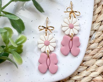 May include: A pair of dangle earrings featuring a pink butterfly charm and a white flower with a pink centre. The earrings have gold accents and are displayed on a white surface.