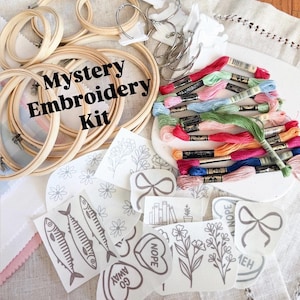 May include: A "Mystery Embroidery Kit" with wooden hoops, embroidery floss in various colors, and printed fabric designs. Designs include flowers, fish, bows, and the words "Nope" and "Go Away".