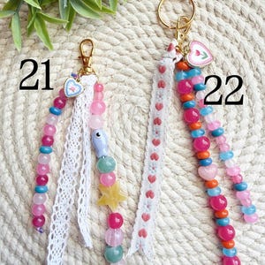 May include: Two beaded keychains with gold-tone hardware. One features pink, blue, and green beads, a star charm, and white lace. The other has pink, orange, and blue beads, a heart charm, and a white ribbon with red hearts. Numbers 21 and 22 are visible.