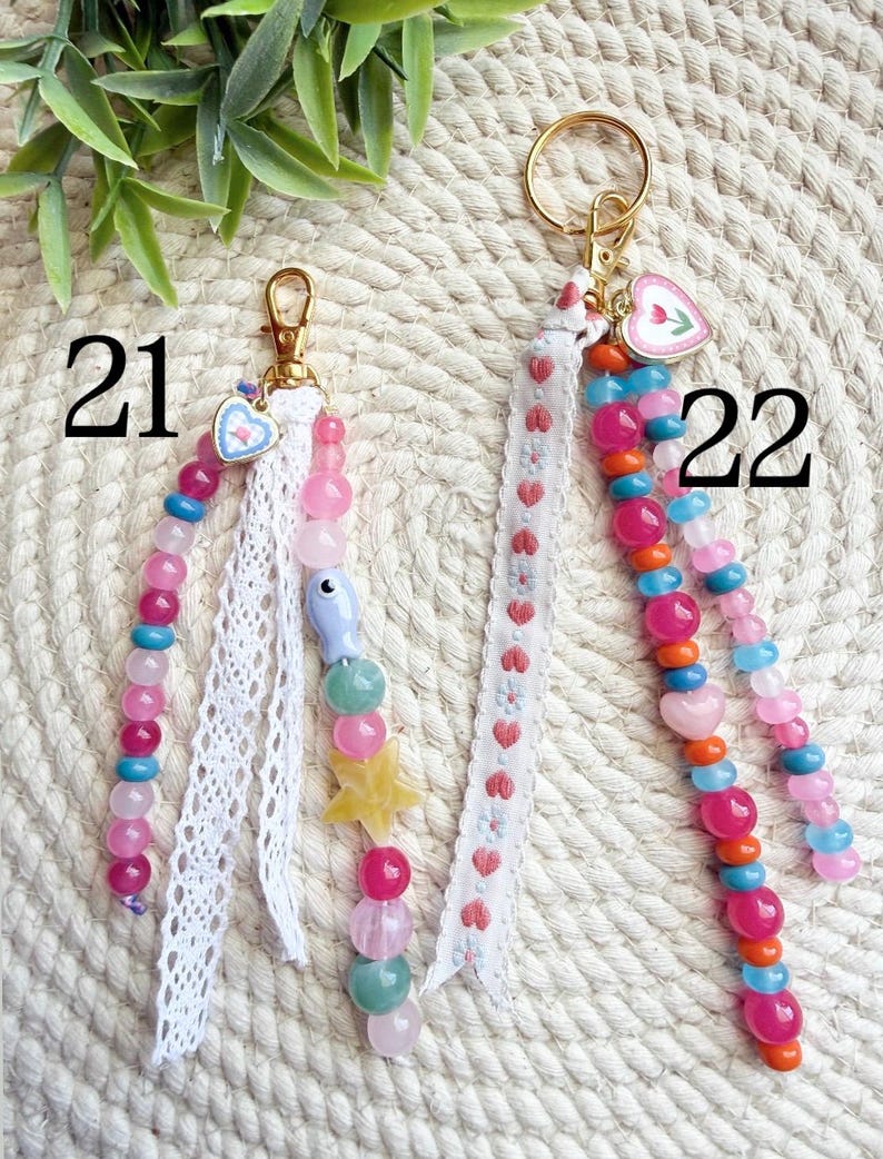 May include: Two beaded keychains with gold-tone hardware. One features pink, blue, and green beads, a star charm, and white lace. The other has pink, orange, and blue beads, a heart charm, and a white ribbon with red hearts. Numbers 21 and 22 are visible.