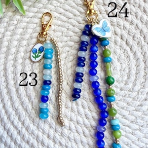 May include: Two beaded keychains with gold-colored clasps. One features a blue floral charm and a strand of blue and white beads. The other has a butterfly heart charm and strands of blue, green, and turquoise beads. Numbers 23 and 24 are visible.