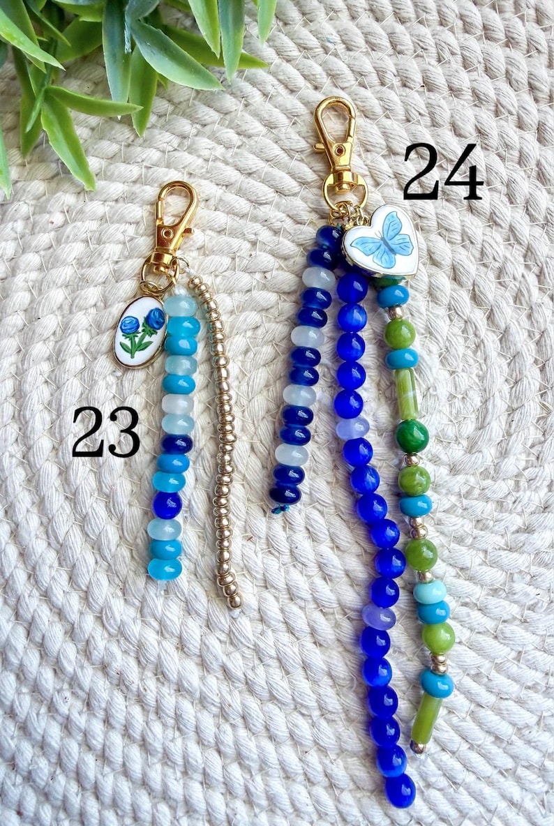 May include: Two beaded keychains with gold-colored clasps. One features a blue floral charm and a strand of blue and white beads. The other has a butterfly heart charm and strands of blue, green, and turquoise beads. Numbers 23 and 24 are visible.