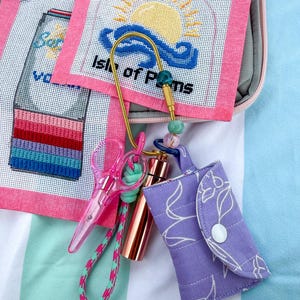May include: A gold carabiner keychain featuring colourful beads, a pair of translucent pink scissors, and a small purple quilted pouch with a white floral outline pattern, displayed alongside two needlepoint canvases with sunset and drink designs.